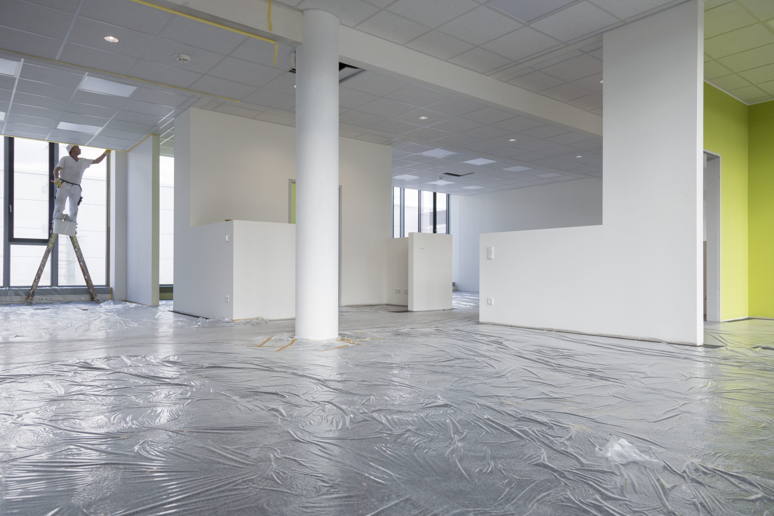 Innenbereich des Bürogebäudes bei den Malerarbeiten, der Fußboden ist. noch mit Folie bedeckt. Links im Hintergrund arbeitet ein Maler an der Wand.