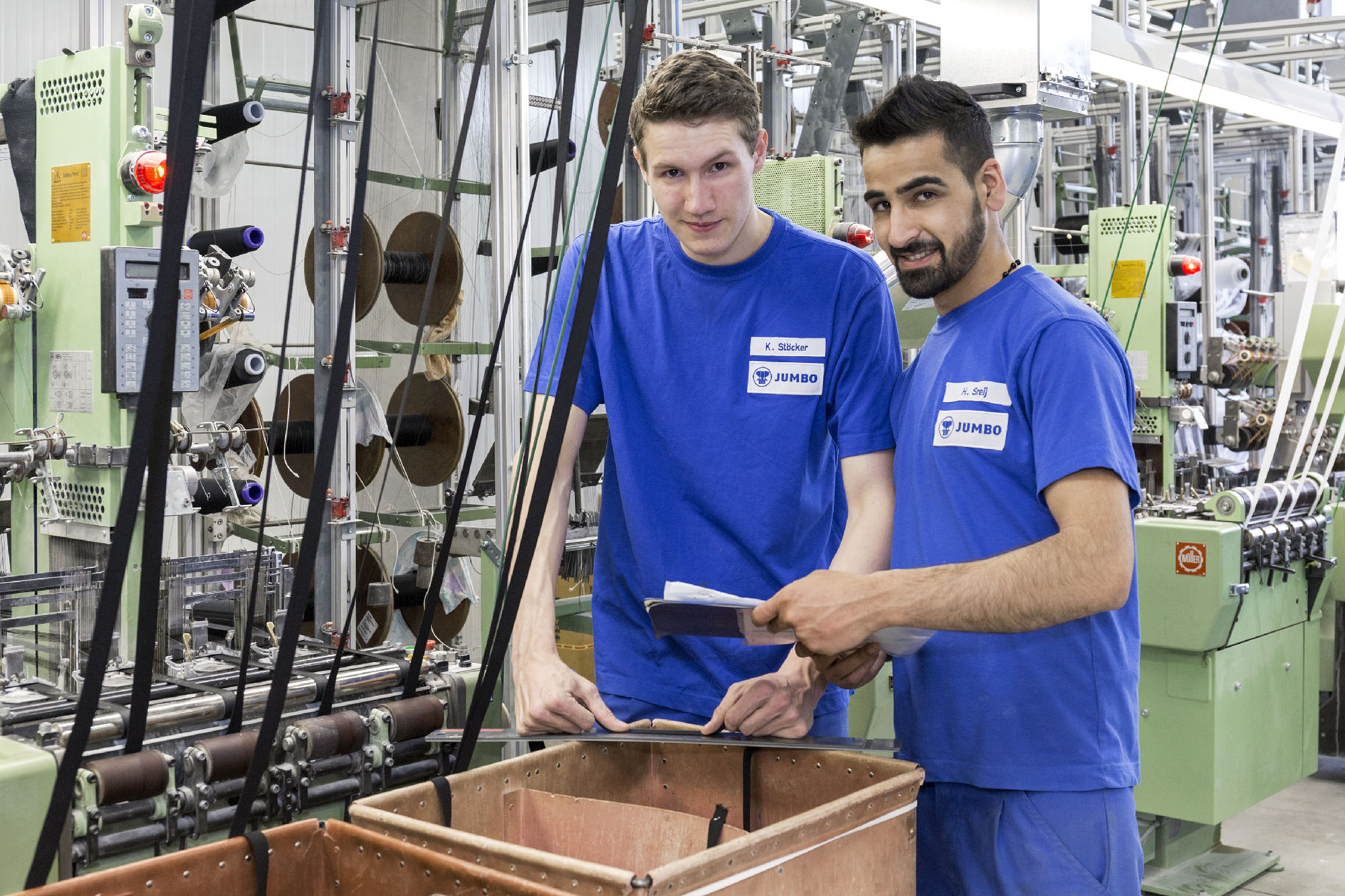 Zwei JUMBO-Textil-Mitarbeiter in der Produktionshalle, sie schauen in die Kamera und tragen blaue Arbeitskleidung