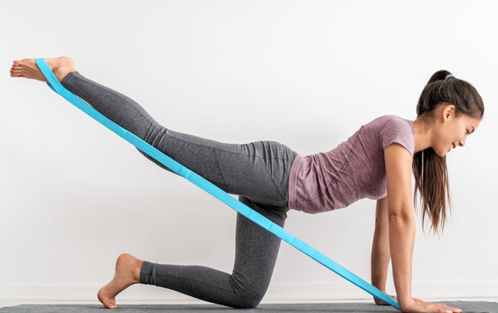 Eine braunhaarige Frau in Yogakleidung kniet auf einer Yogamatte. Sie trainiert mit einem elastischen Fitnesstextil in türkis ihre Beinmuskulatur, indem sie das Band mit einem Bein nach hinten streckt und vorne festhält.
