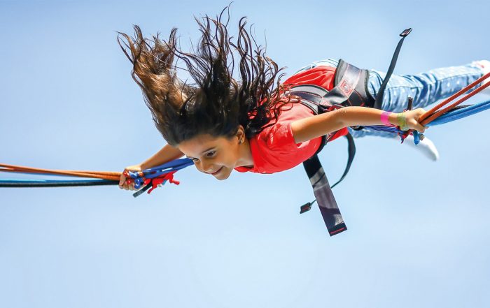 Ein braunhaariges Mädchen befindet sich gerade in der Luft, die Haare fliegen nach oben. Sie hängt in einem Sichtheitsgurts eines Trampolins.