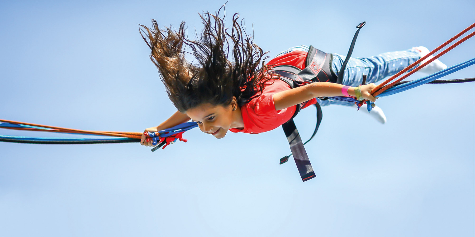 Ein braunhaariges Mädchen befindet sich gerade in der Luft, die Haare fliegen nach oben. Sie hängt in einem Sichtheitsgurts eines Trampolins.