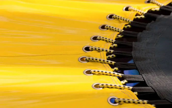 Eine Nahaufnahme eines Trampolins. Die linke Bildhälfte zeigt den gelben Außenrand aus Stoff, die rechte Bildhälfte den Anschnitt der schwarzen Sprungfläche. Mittig sind die schwarz-gelben Gummibänder zu sehen, die beides miteinander verbinden.