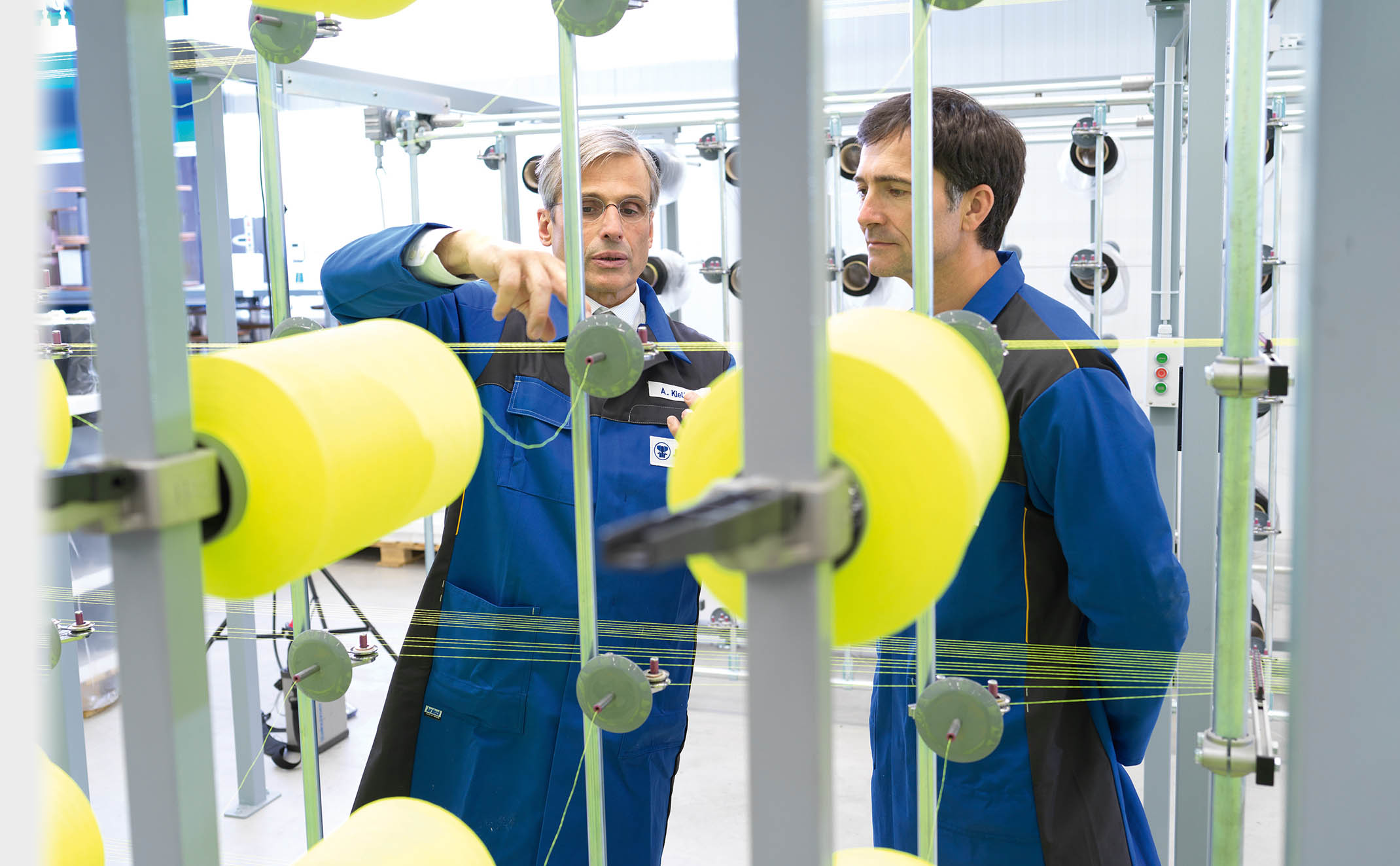 Zwei JUMBO-Textil-Mitarbeiter in blauer Arbeitskleidung hinter einer Maschine in der Produktion