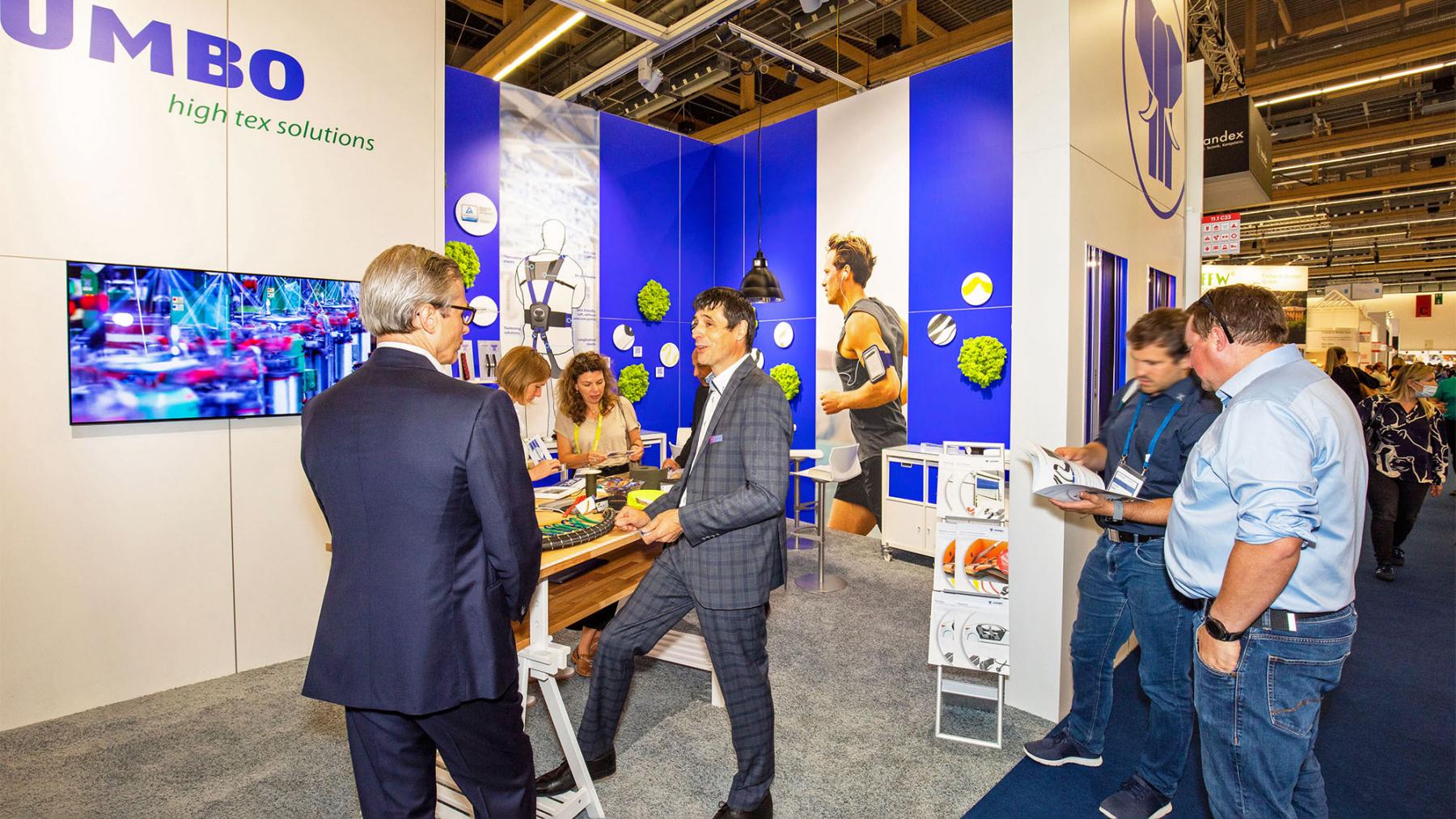 JUMBO-Textil-Innovationen auf der techtextil 2022: Zwei JUMBO-Textil Mitarbeiter im Gespräch miteinander, im Hintergrund der Messestand in blau und weiß, rechts betrachten zwei Messebesucher eine Broschüre