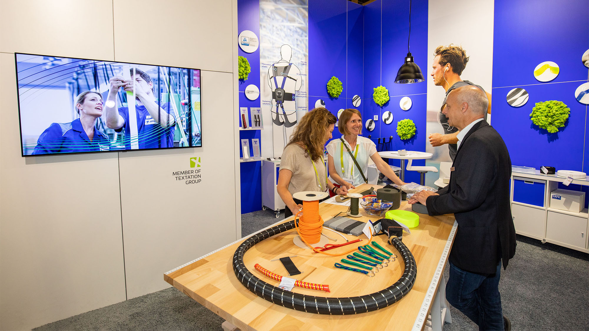 JUMBO-Textil-Innovationen auf der techtextil 2022: Ein Mitarbeiter von JUMBO-Textil berät zwei Frauen an einem Tisch mit verschiedenen Produktbeispielen. Links ist ein Bildschirm, auf dem gerade zwei weitere Mitarbeiter zu sehen sind (Standbild aus dem Imagevideo).