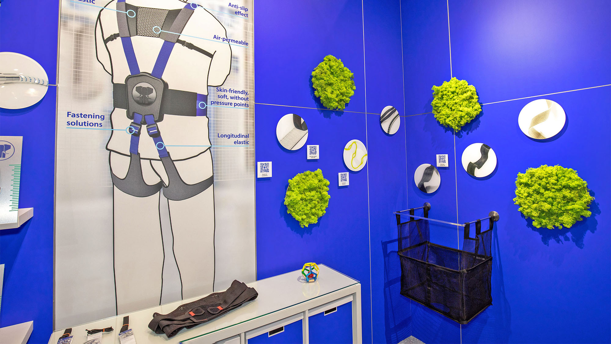 Messestand von JUMBO-Textil im Detail: blaue Messewand, darauf Produktbilder verschiedene Schmaltextilien, im Vordergrund sind Produktbeispiele ausgestellt. Links befindet sich eine Grafik eines Exoskeletts