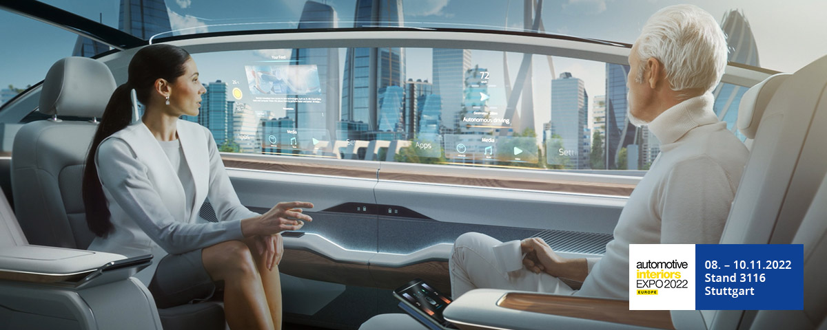 JUMBO-Textil auf der AIE 2022: Eine dunkelhaarige Frau und ein weißhaariger Mann sitzen sich in einem Auto gegenüber. Das Auto hat eine futuristische Inneneinrichtung und sie fahren an einer ebenfalls futuristischen Großstadtkulisse vorbei. Im Fenster sind zwei Projektionsdisplay-Anzeigen zu sehen. Unten rechts befindet sich ein Hinweis auf die Automotive Interiors Expo 2022.