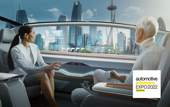 Eine dunkelhaarige Frau und ein weißhaariger Mann sitzen sich in einem Auto gegenüber. Das Auto hat eine futuristische Inneneinrichtung und sie fahren an einer ebenfalls futuristischen Großstadtkulisse vorbei. Im Fenster sind zwei Projektionsdisplay-Anzeigen zu sehen. Unten rechts befindet sich ein Hinweis auf die Automotive Interiors Expo 2022.