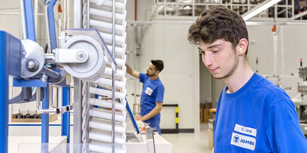 Ein Produktionsmitarbeiter mit blauem Jumbo-Textil-Shirt an einer Maschine in einer Produktionshalle, ein weiterer Mitarbeiter im Hintergrund.