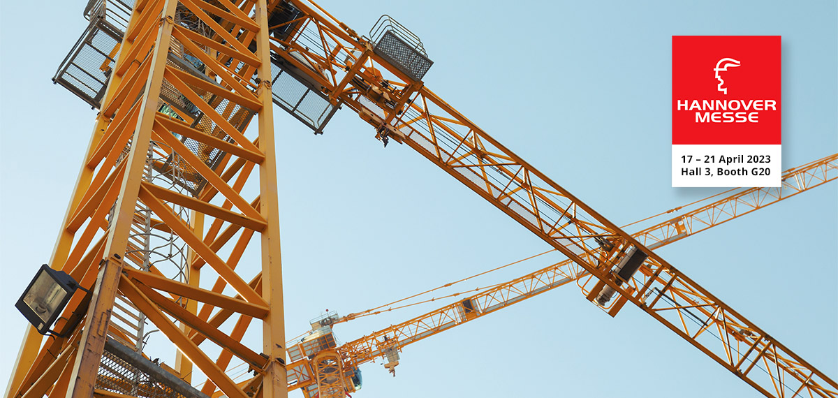Landscape photo showing two orange cranes. The perspective is from the bottom up, background is a cloudless blue sky. One crane is in the foreground, the other in the background. On the right you can see the logo of Hannover Messe.