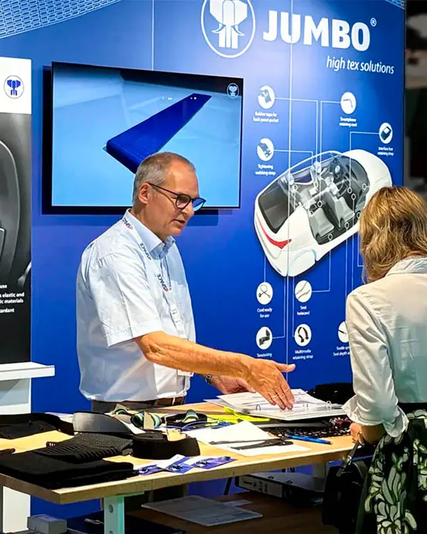 Mitarbeiter am Messestand im Gespräch mit einer Kundin, im Hintergrund ein Bildschirm mit Produktpräsentation und ein Auto-Modell